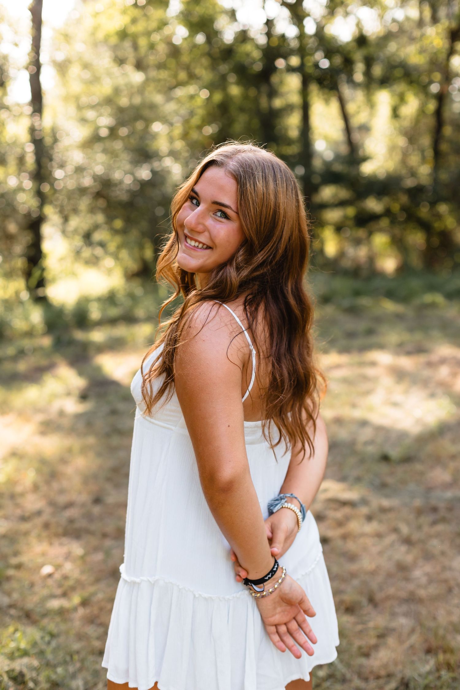 Smiling young woman in a white sundress with bracelets posing in a sunlit forest setting.