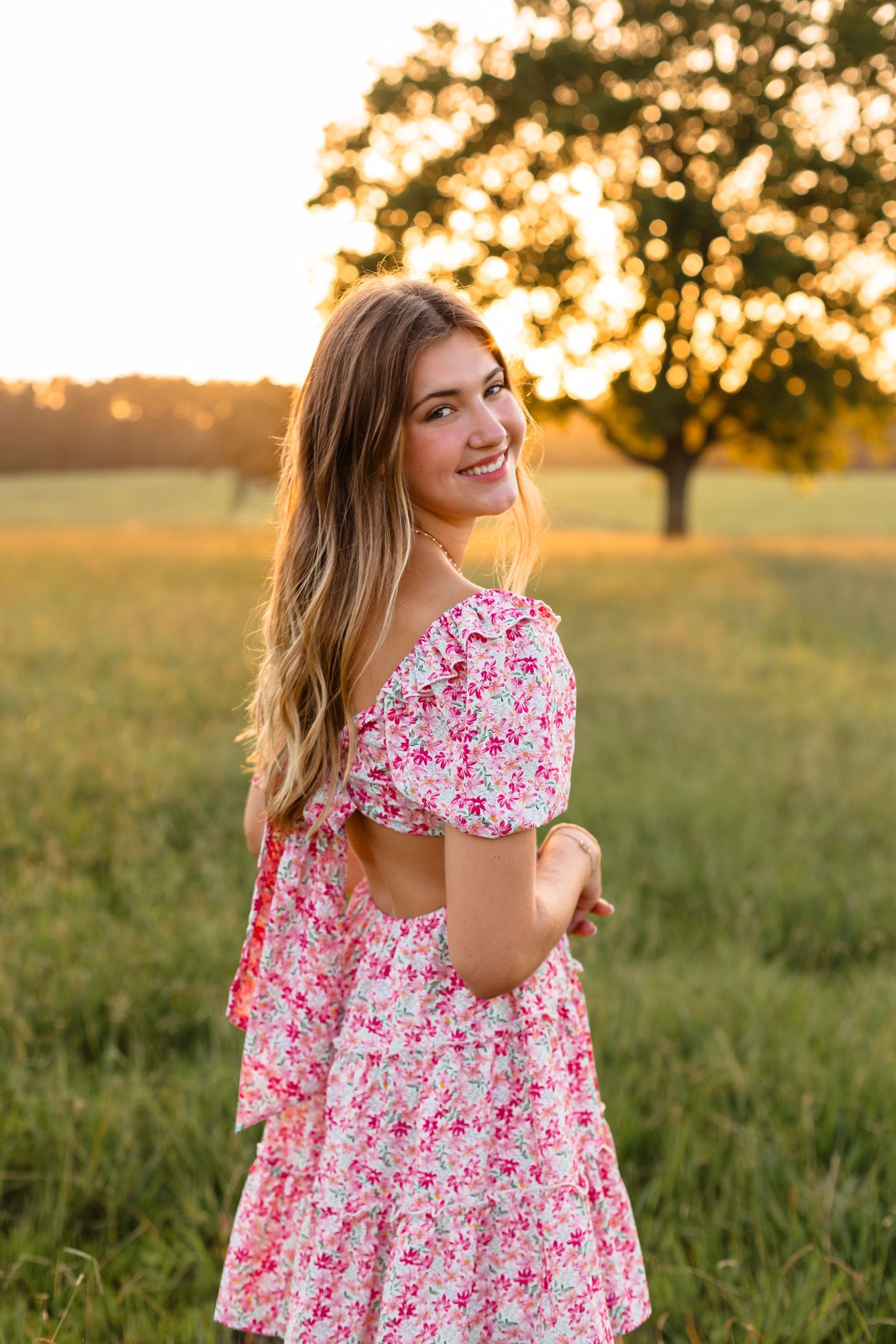 Smiling young woman in a pink floral off-shoulder dress posing in a golden sunset field.
