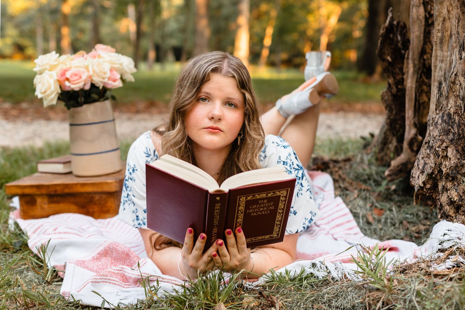 Young girl lying on a blanket outdoors reading a book near a tree with pink flowers.