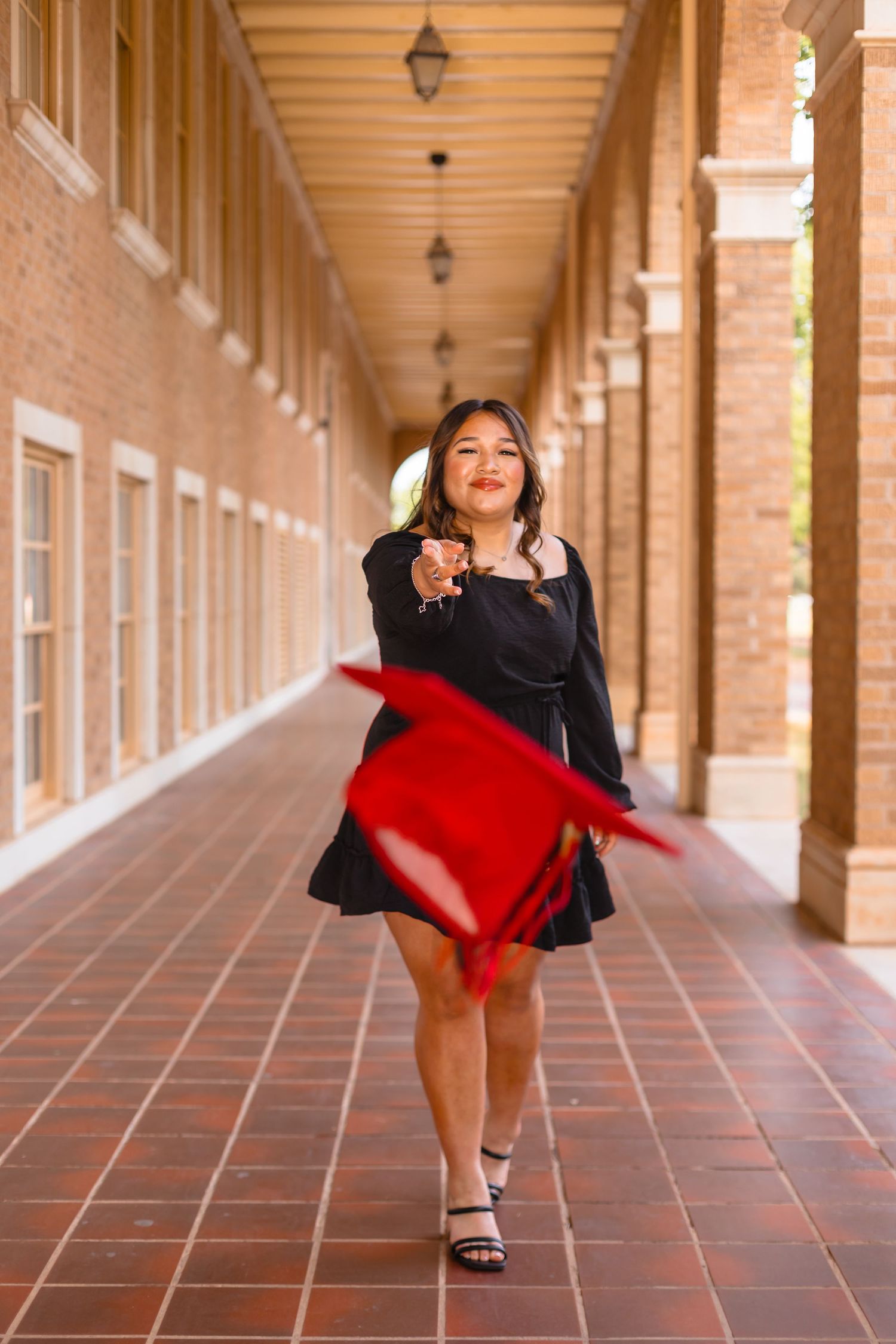 Young woman in black dress tossing red graduation cap in brick university hallway, celebrating academic achievement.