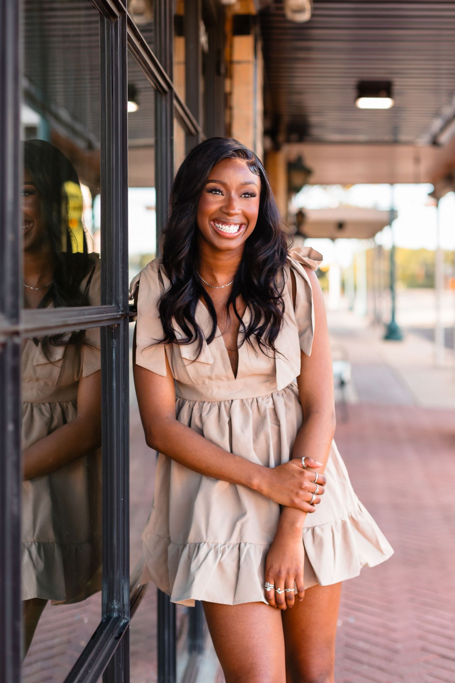 Smiling young Black woman in a tan mini dress leaning against a glass storefront on a sidewalk.