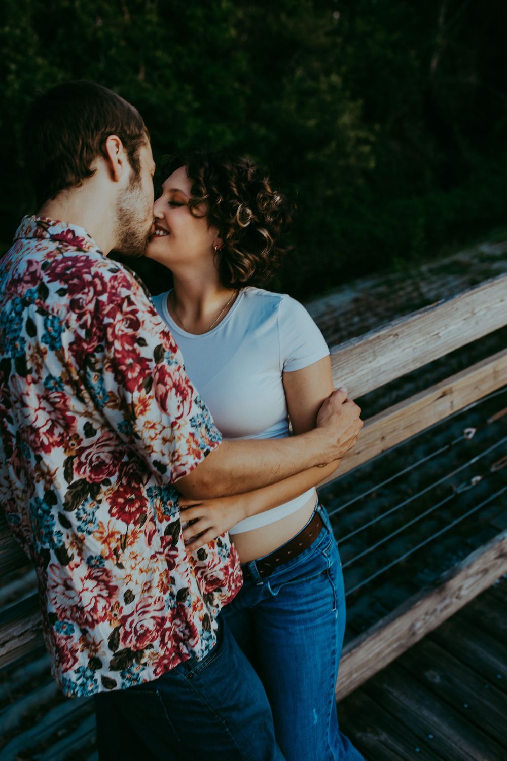A couple embraces intimately on a wooden dock surrounded by green foliage on a summer day.