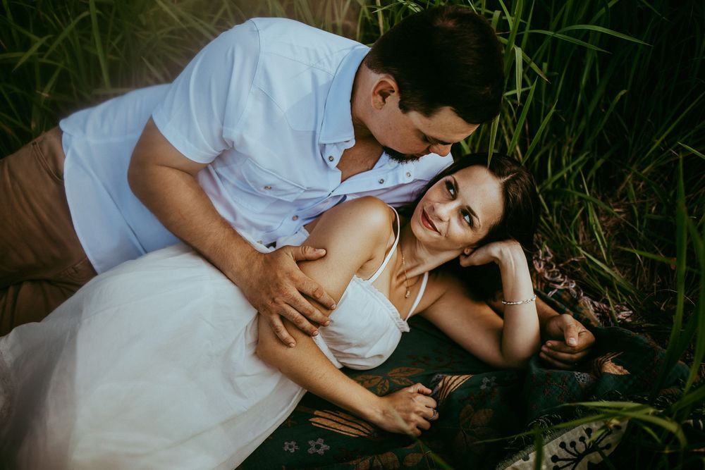 A romantically posed couple wearing white clothing lay together in green grass during an outdoor photo session.
