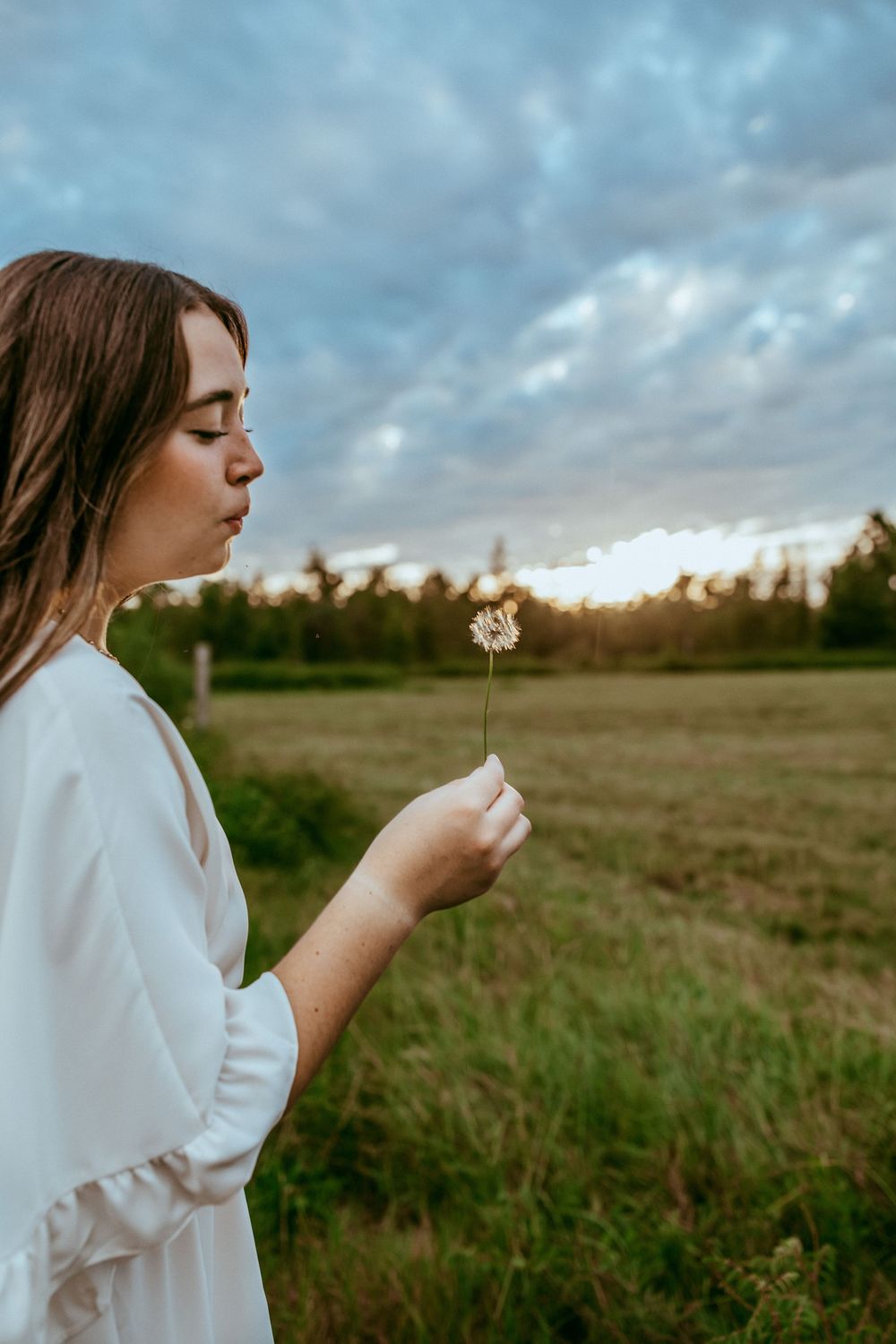 A woman in white stands in a field at dusk gazing at a delicate dandelion seed head under a cloudy sky.