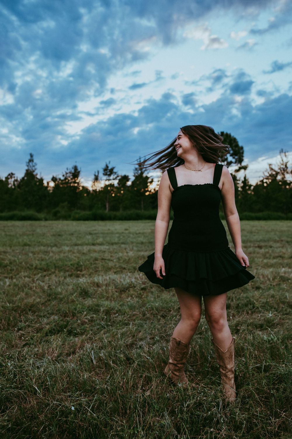 Person in a black dress twirling in an open field under dramatic cloudy skies at dusk.