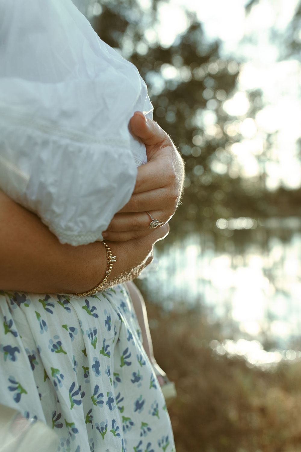 Close-up photo of a pregnant belly in a floral dress against a blurry outdoor background with water reflections.