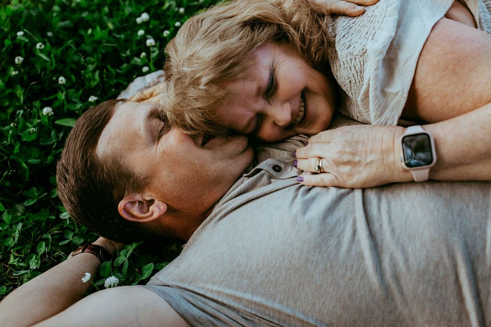 A couple lies together on green grass, sharing an intimate moment outdoors on a sunny day.