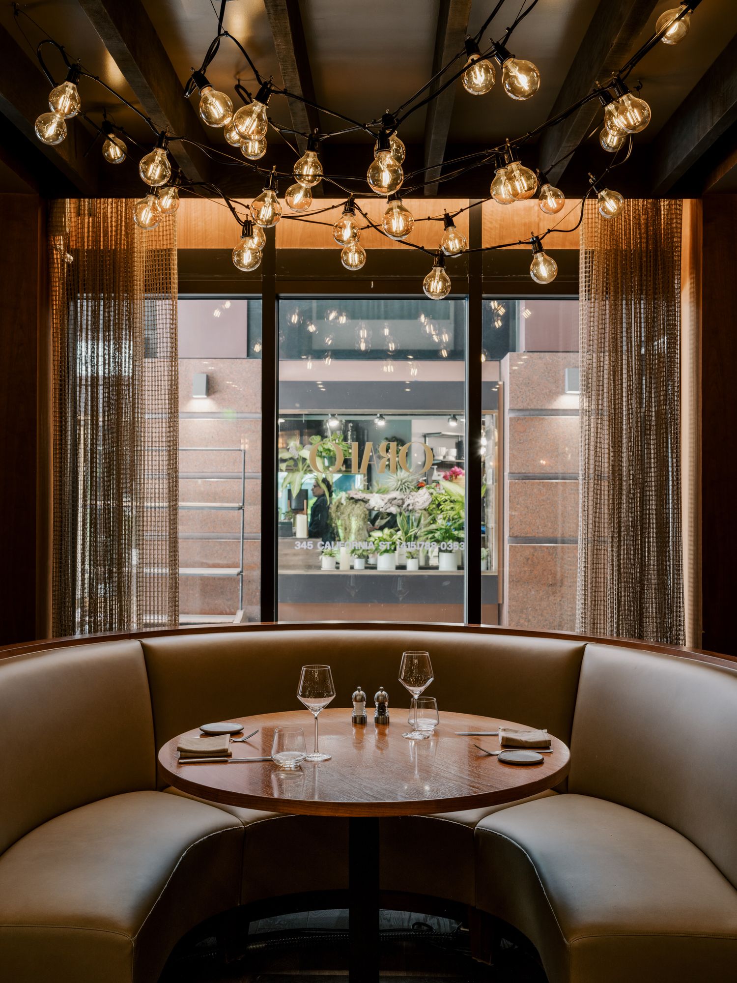 Intimate restaurant booth with curved brown leather seating below string lights and window view of retail display.