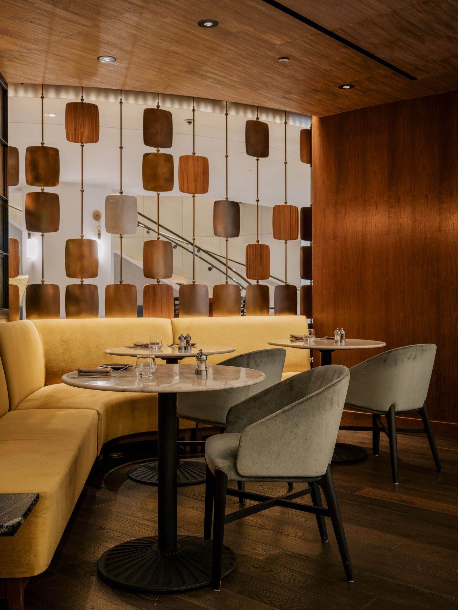 Modern restaurant interior with yellow banquette seating, grey chairs and geometric wall art against wood paneled walls.