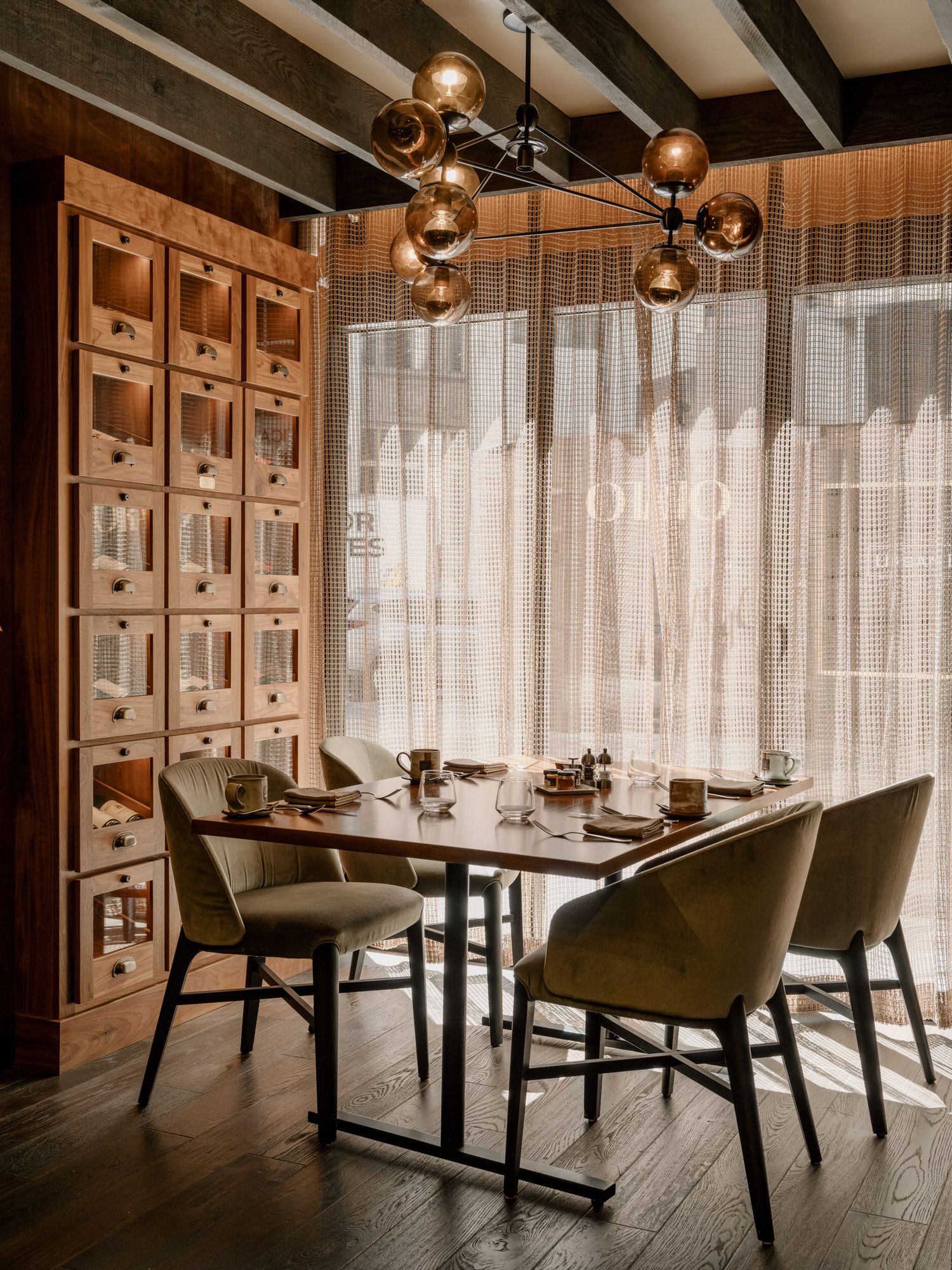 A cozy restaurant dining area with modern pendant lights, olive green chairs, and sheer curtains filtering natural light.