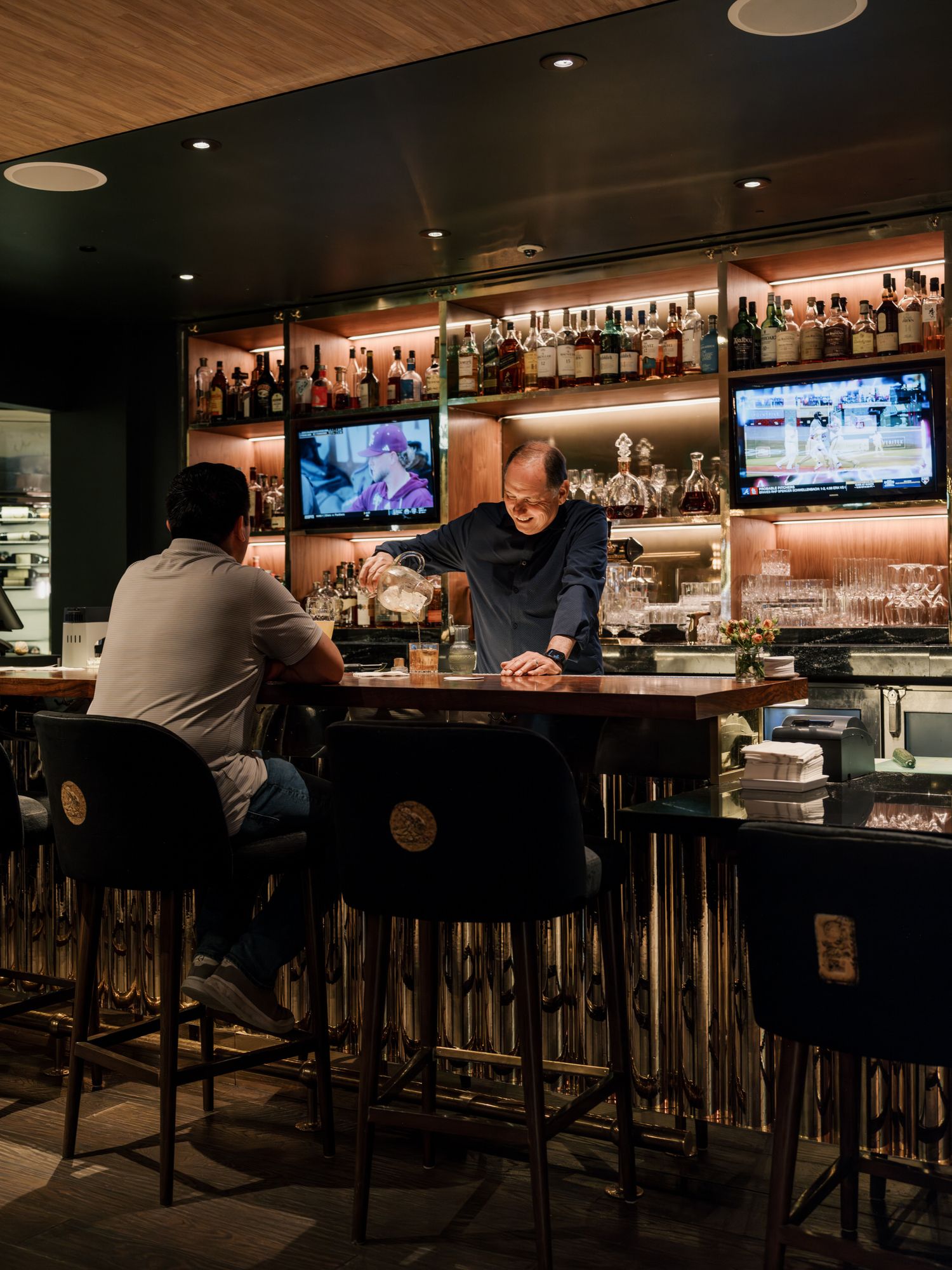 Evening scene at a dimly lit bar counter with patrons seated on barstools and TVs mounted above shelves of liquor.