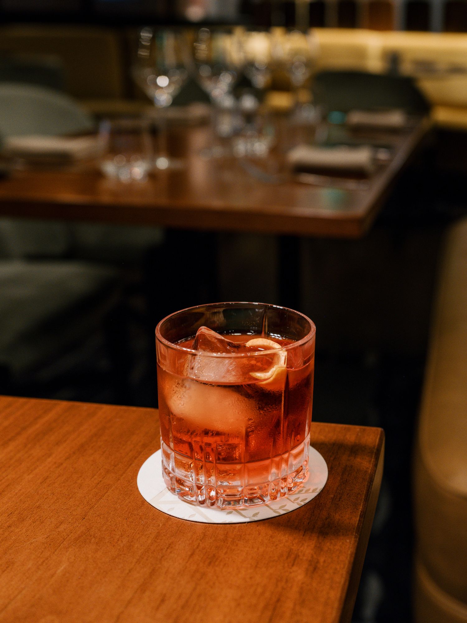 A classic cocktail with orange garnish sits on a crystal glass with large ice cube on a wooden bar table.