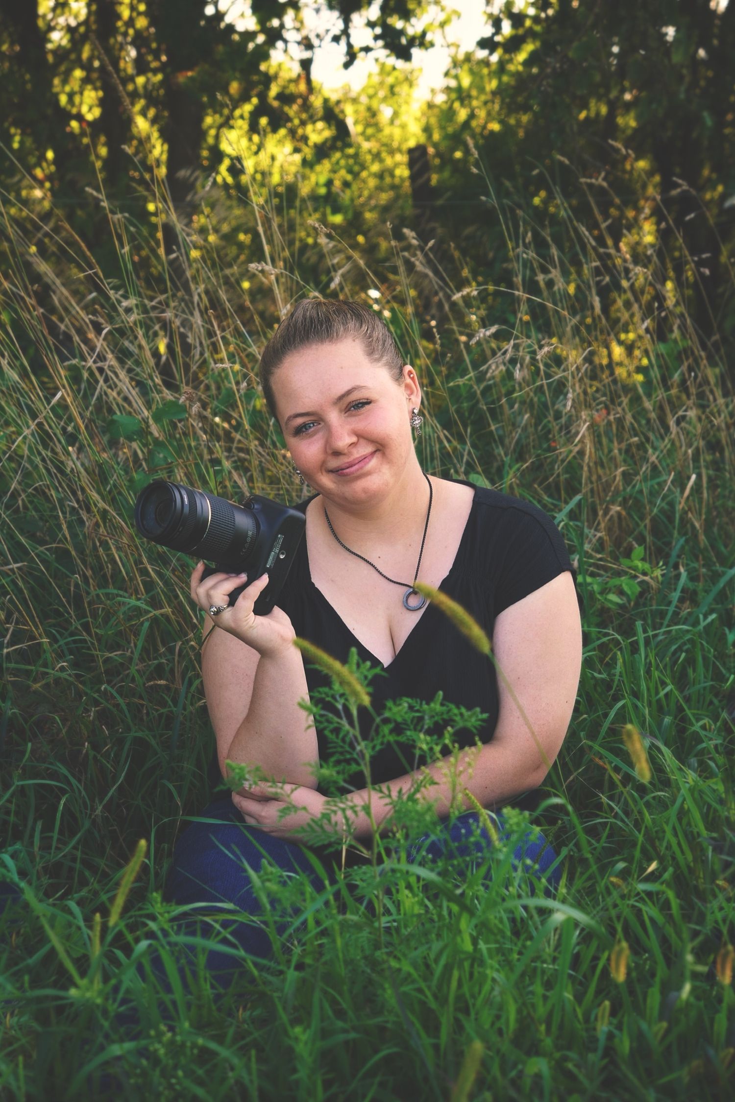 A photographer in a black top poses with their camera while sitting in tall grass at sunset.