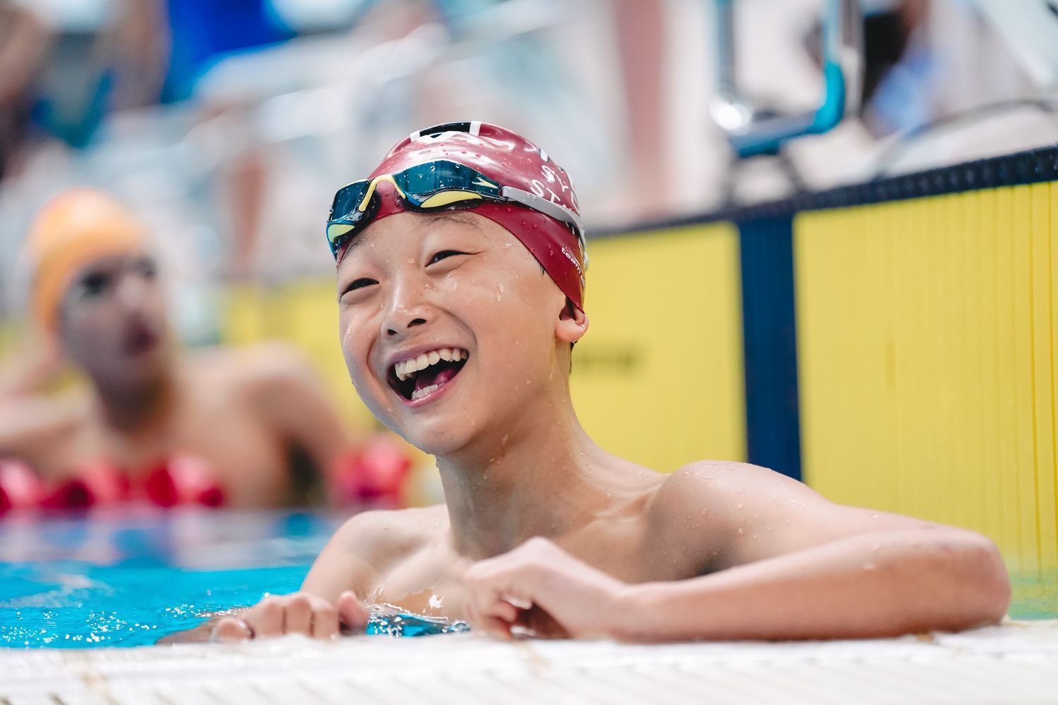 Swimmer in red cap and goggles laughing joyfully at pool's edge.