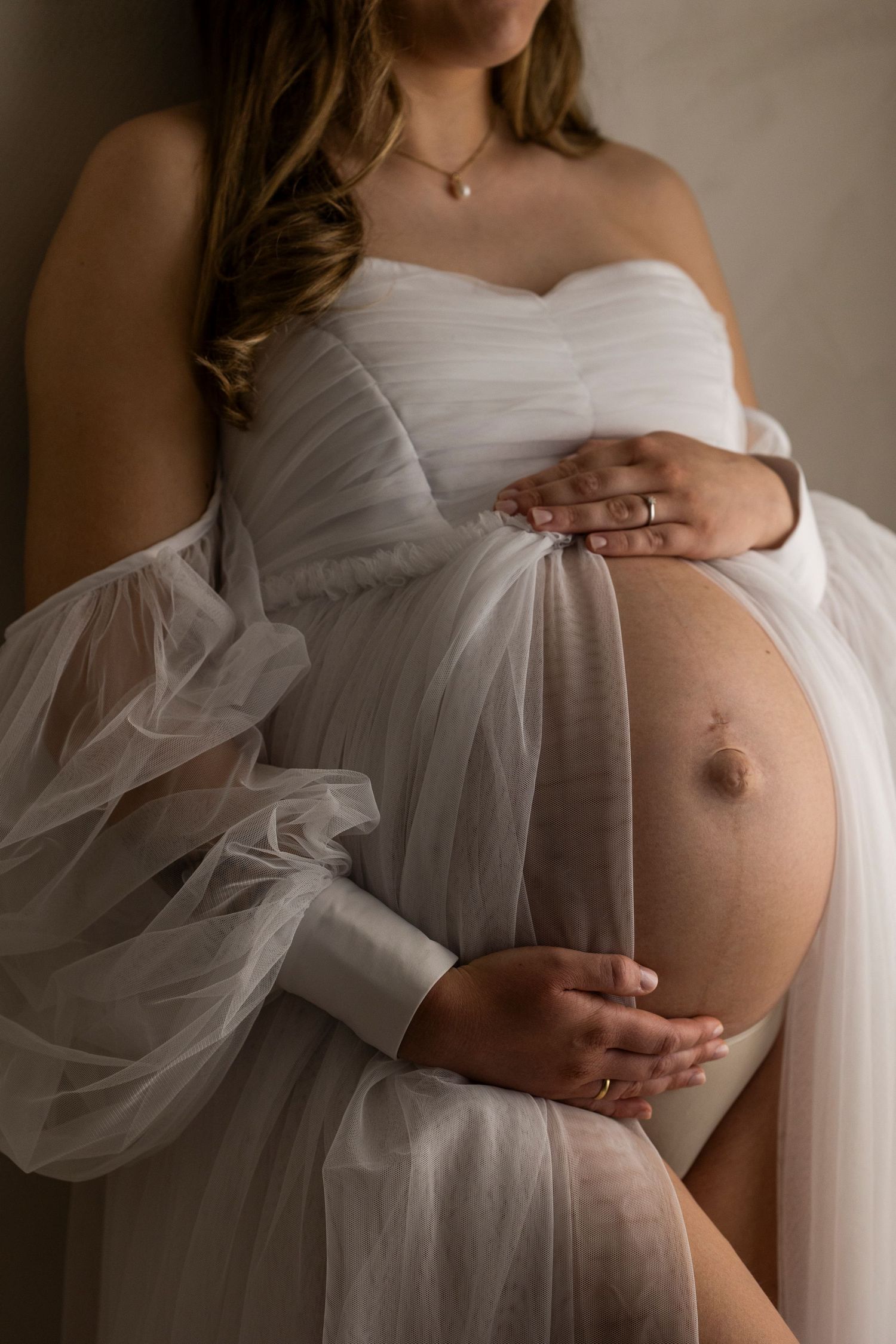 Close-up of pregnant belly in white maternity dress with hand wearing diamond engagement ring resting on bump.