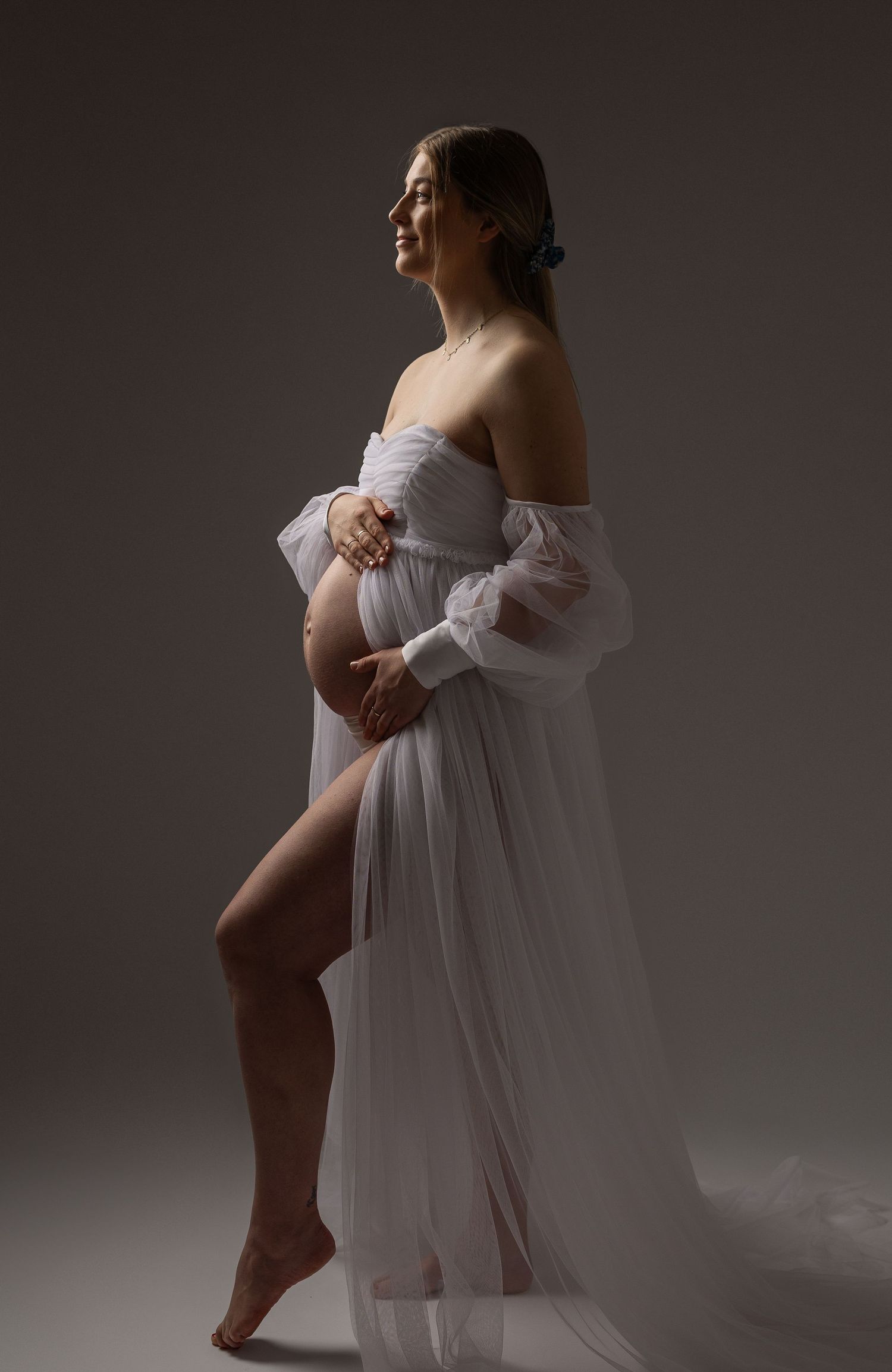 Close-up of pregnant belly in white maternity dress with hand wearing diamond engagement ring resting on bump.