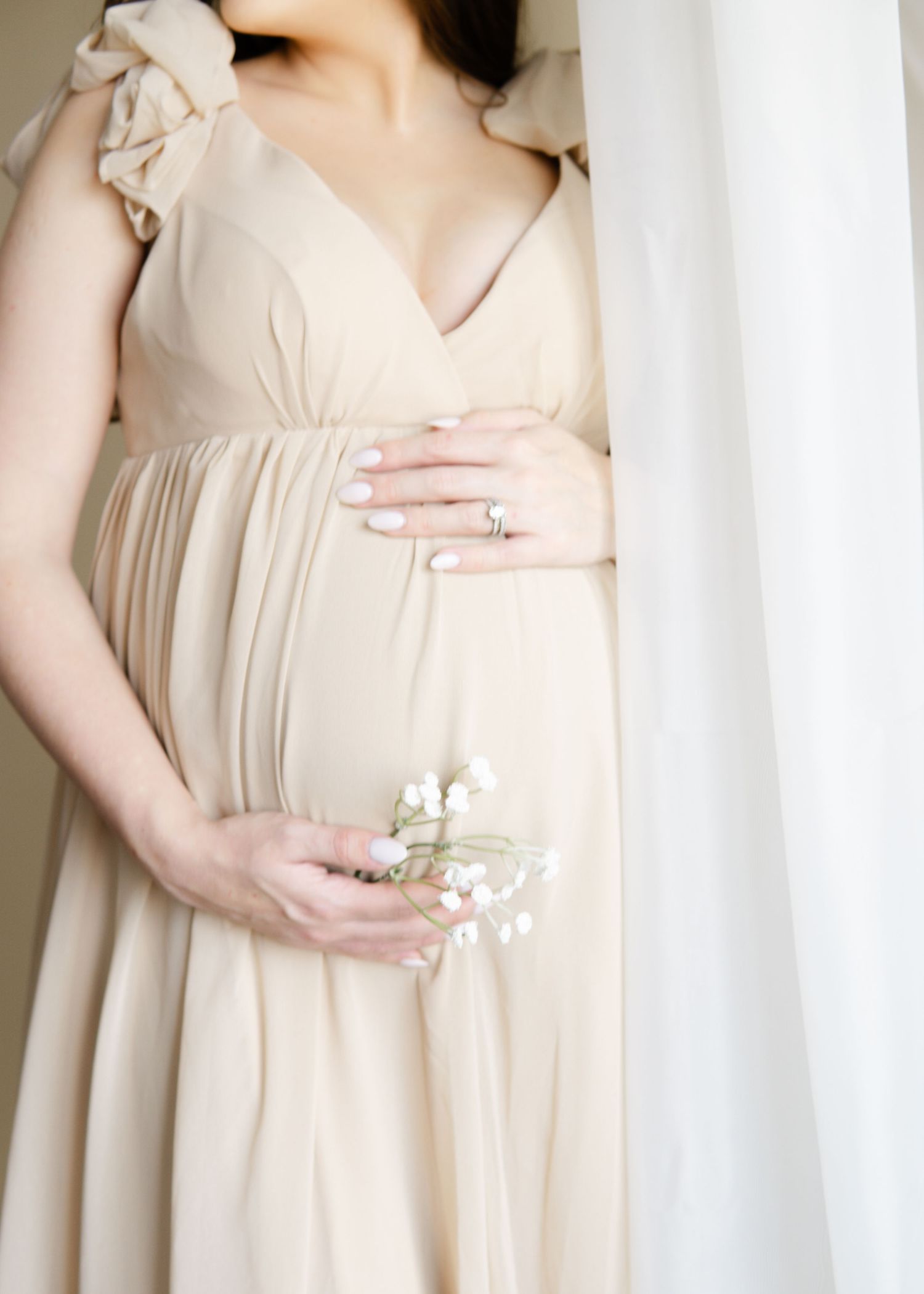 Maternity detail shot, expecting mother’s hands resting on belly, beige floral dress with ruffles