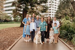 Large multigenerational family posing with golden retriever on wooden path in lush park with city buildings.