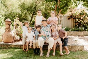 Elderly man sitting on a bench surrounded by seven grandchildren in a lush garden during a family portrait.