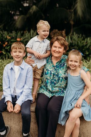 A smiling woman poses with three young children on a stone bench surrounded by tropical greenery.