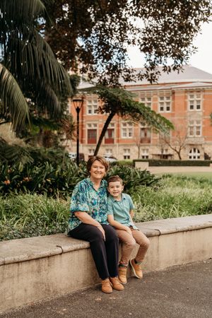 A woman and young boy sit together on a stone wall in a park, with a red brick building and palm tree behind them.