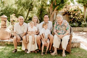 Multi-generational family of six smiling together on a wicker bench outdoors in a sunny garden setting.