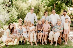 Large multigenerational family portrait outdoors in lush garden setting, smiling group of adults and children in neutral tones.