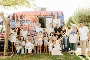 Large group of adults and children posing in front of a colorful soft serve ice cream food truck outdoors.