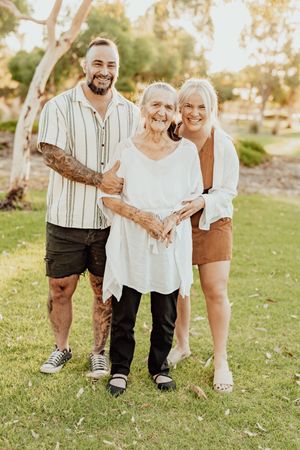 Three generations of family smiling together outdoors in a sunny park, including a tattooed young man and two women.