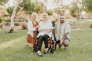 Two women and a man pose outdoors in a park, one woman seated in a wheelchair, smiling together.