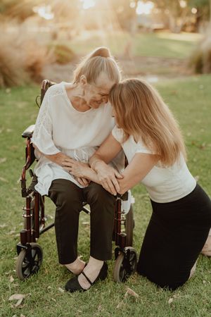 Elderly woman in wheelchair shares tender moment with younger woman kneeling beside her in sunlit park.