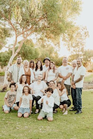 Large multigenerational family group photo outdoors on green grass under trees, wearing coordinated white and neutral outfits.