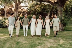 Seven people holding hands walking together on green grass under large trees in a sunny outdoor family portrait.