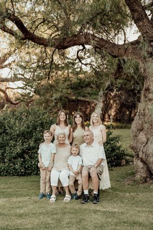 Multi-generational family portrait outdoors under large tree, grandparents seated with grandchildren and adult daughters standing behind.