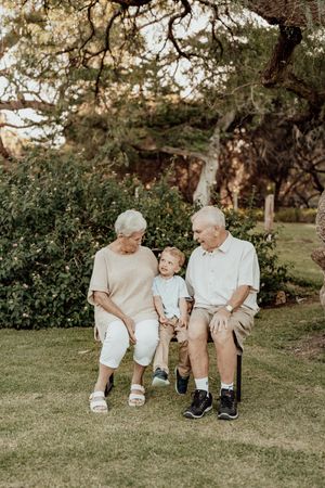 Elderly couple sitting on a bench outdoors with a toddler between them, surrounded by lush green trees.