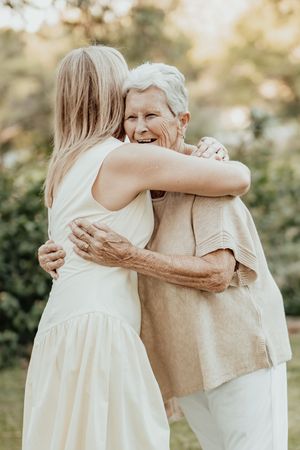Two women sharing a warm embrace outdoors, younger blonde woman in white dress hugging smiling elderly woman in beige top.