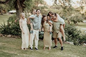 A joyful group of six friends posing outdoors in neutral tones, laughing and giving piggyback rides in a lush garden setting.