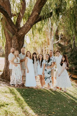 A group of people in coordinated light-colored outfits pose together under willow trees in a sunlit outdoor setting.