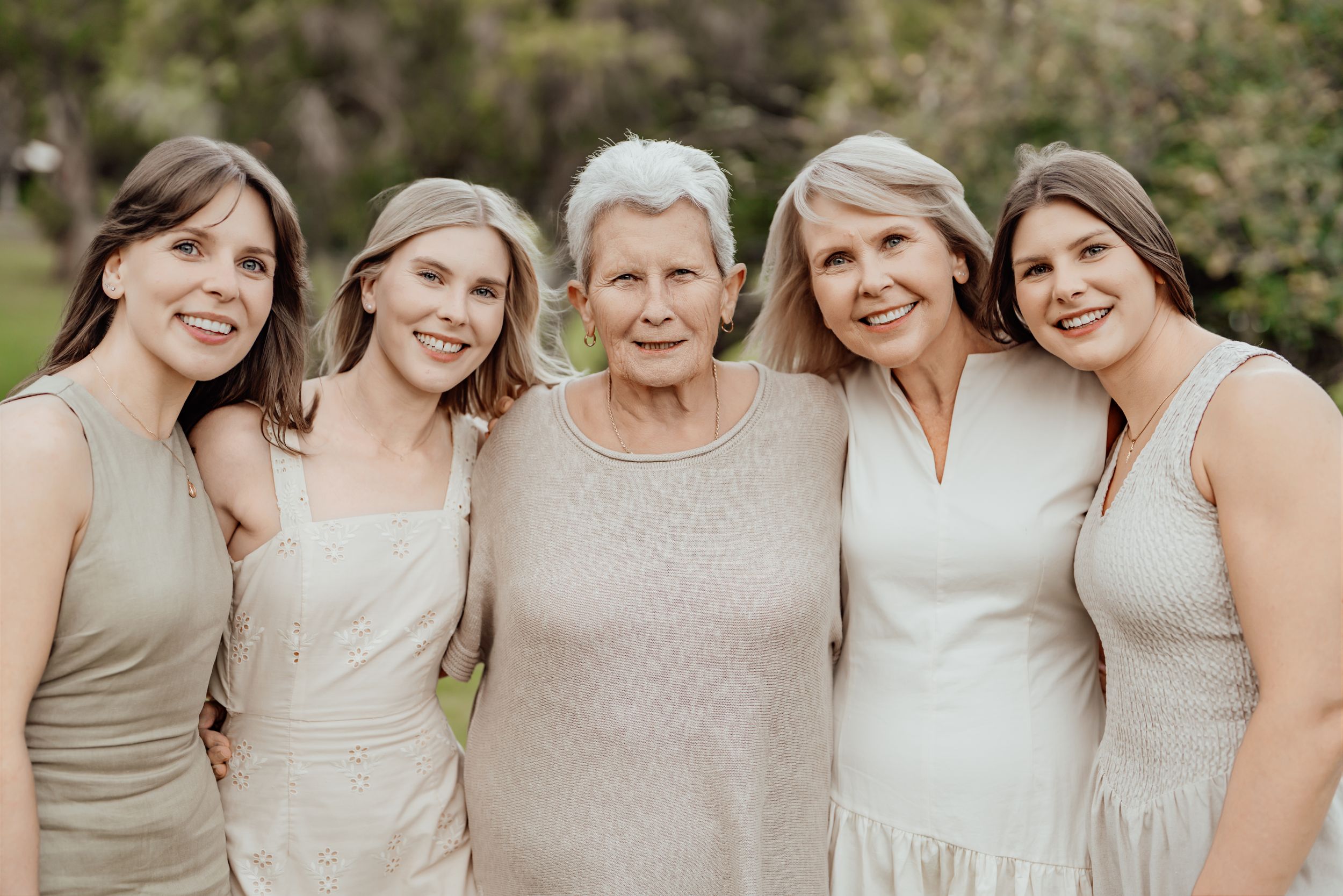 Five women of different ages pose together outdoors, smiling warmly in coordinating neutral-toned outfits.