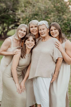 Five women of different generations pose together outdoors in neutral tones, smiling warmly in a lush garden setting.