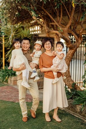 Happy Asian family of five posing outdoors, parents holding three young children near a tree in a garden.