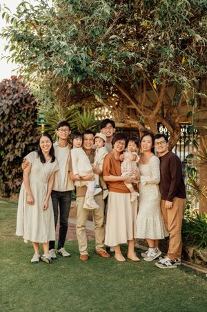 A large Asian family posing together outdoors in a lush garden, wearing coordinated neutral and earth-toned outfits.