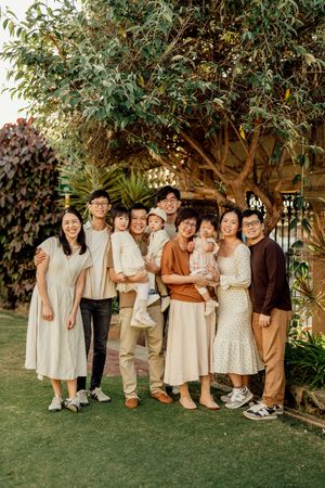 Large Asian family posing together outdoors in a lush garden, wearing neutral tones, including children and a baby.