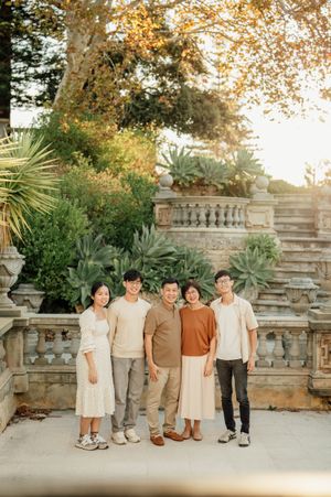A family of five poses together in front of a stone balustrade and lush garden, dressed in neutral tones.