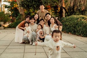 Joyful Asian family group photo outdoors, laughing together while a toddler runs toward camera in a garden setting.
