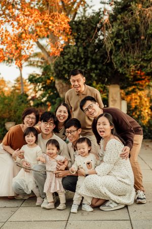Large Asian family posing together outdoors in autumn, smiling with young children, surrounded by colorful fall foliage.
