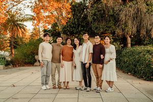 A joyful Asian family of seven poses together outdoors among autumn trees on a stone patio.
