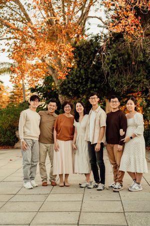 A family of seven poses together outdoors among autumn foliage, dressed in warm neutral tones.