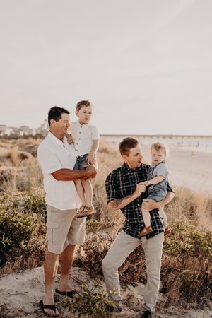 Two adults holding toddlers pose on coastal dunes during sunset.