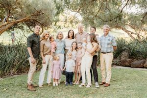 Large family group portrait outdoors on green grass surrounded by trees, featuring adults and young children in coordinated pastel outfits.